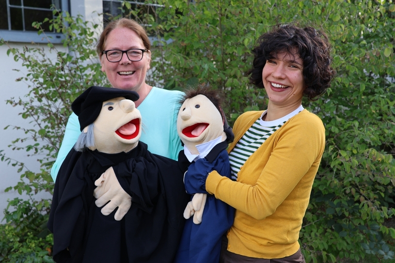 Diakonin Tanja Götz und Pfarrerin Annika Landt mit zwei Handpuppen vor der Pauluskirche.
